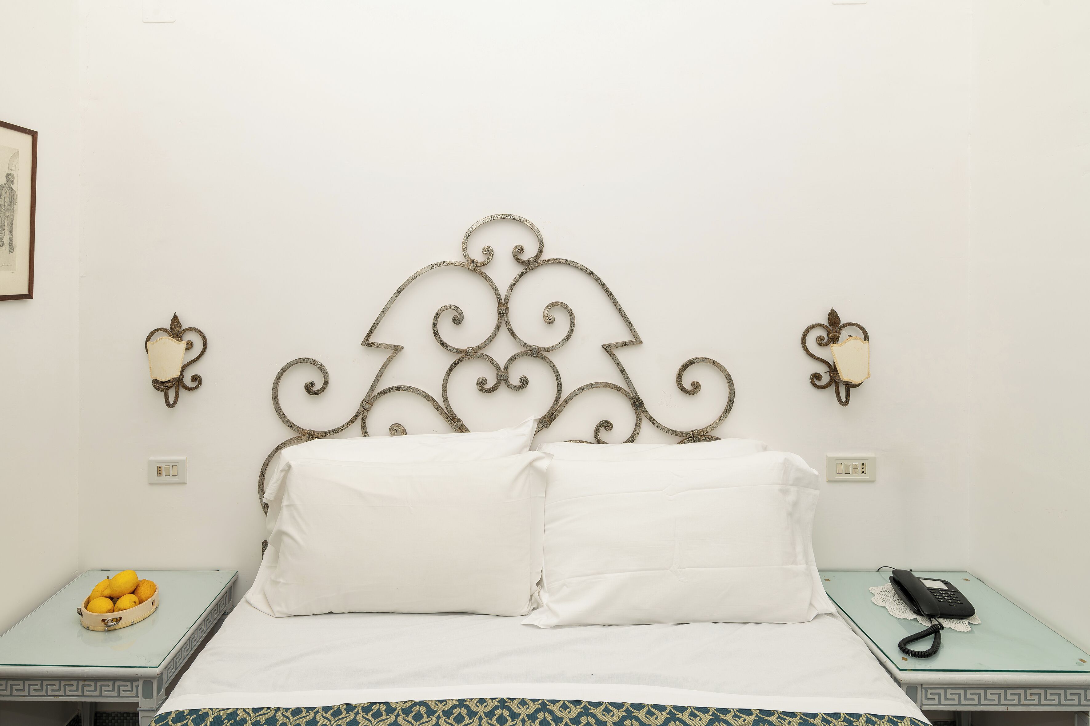 Una cama con ropa blanca, cabecero metálico con detalles decorativos en forma de volutas, mesita de noche con un teléfono y un cuenco de fruta, y lámparas fijadas a la pared.