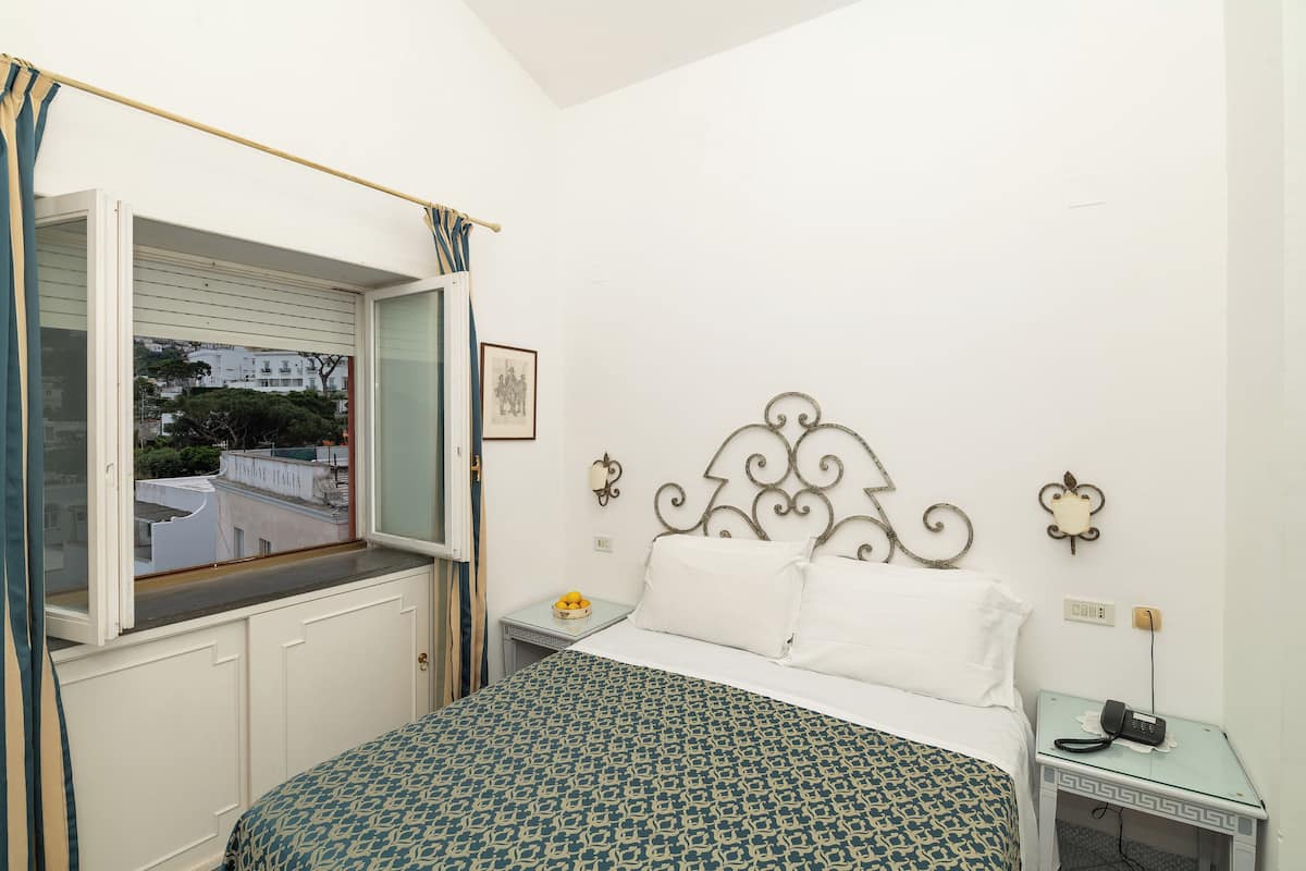 Un dormitorio con cama, ventana con cortinas, mesita de noche con lámpara y teléfono instalado en la pared.
