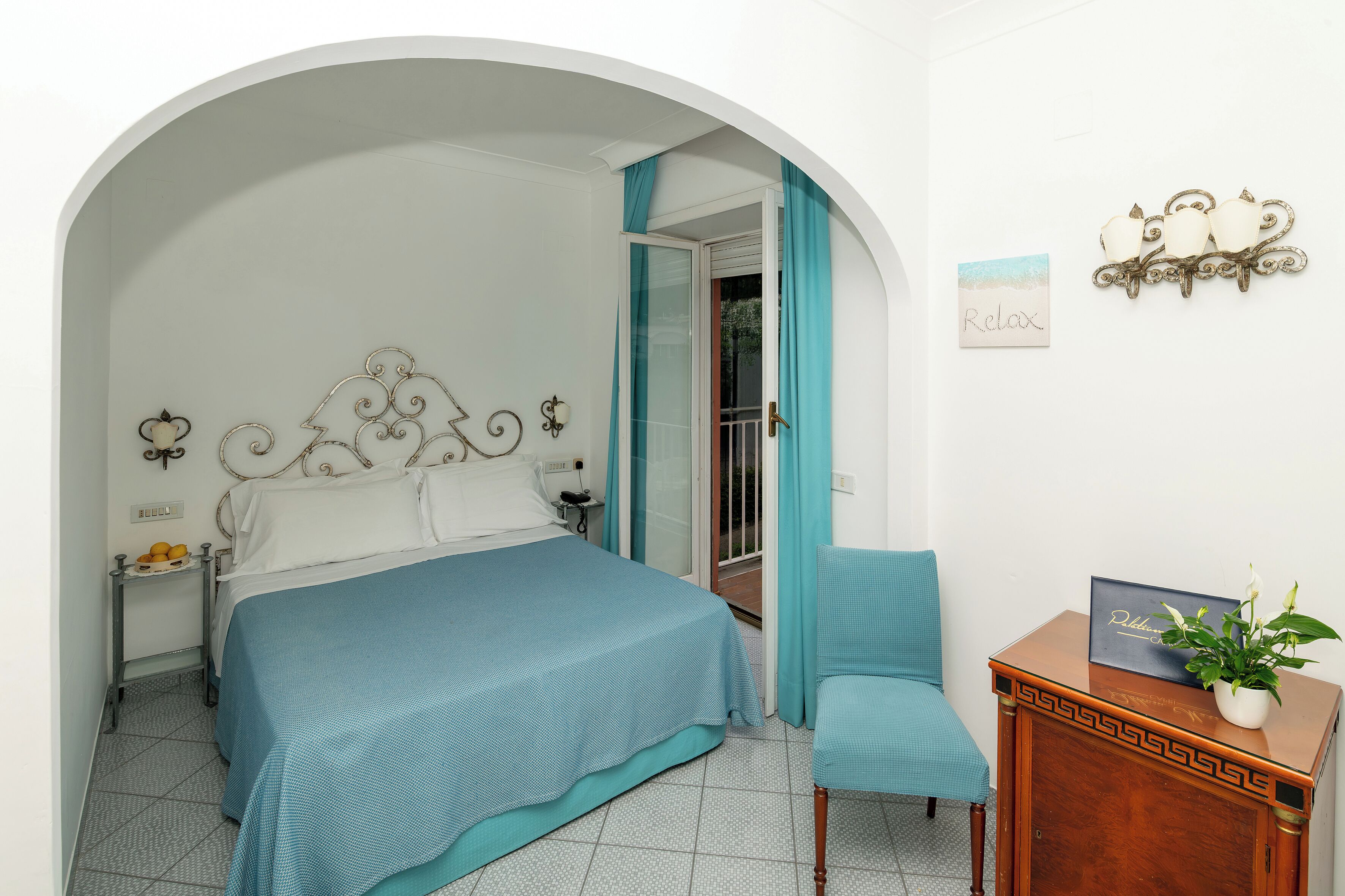 A bedroom with a bed, a chair, a nightstand, a bedside table, a wall-mounted lamp, and a decorative wall piece.