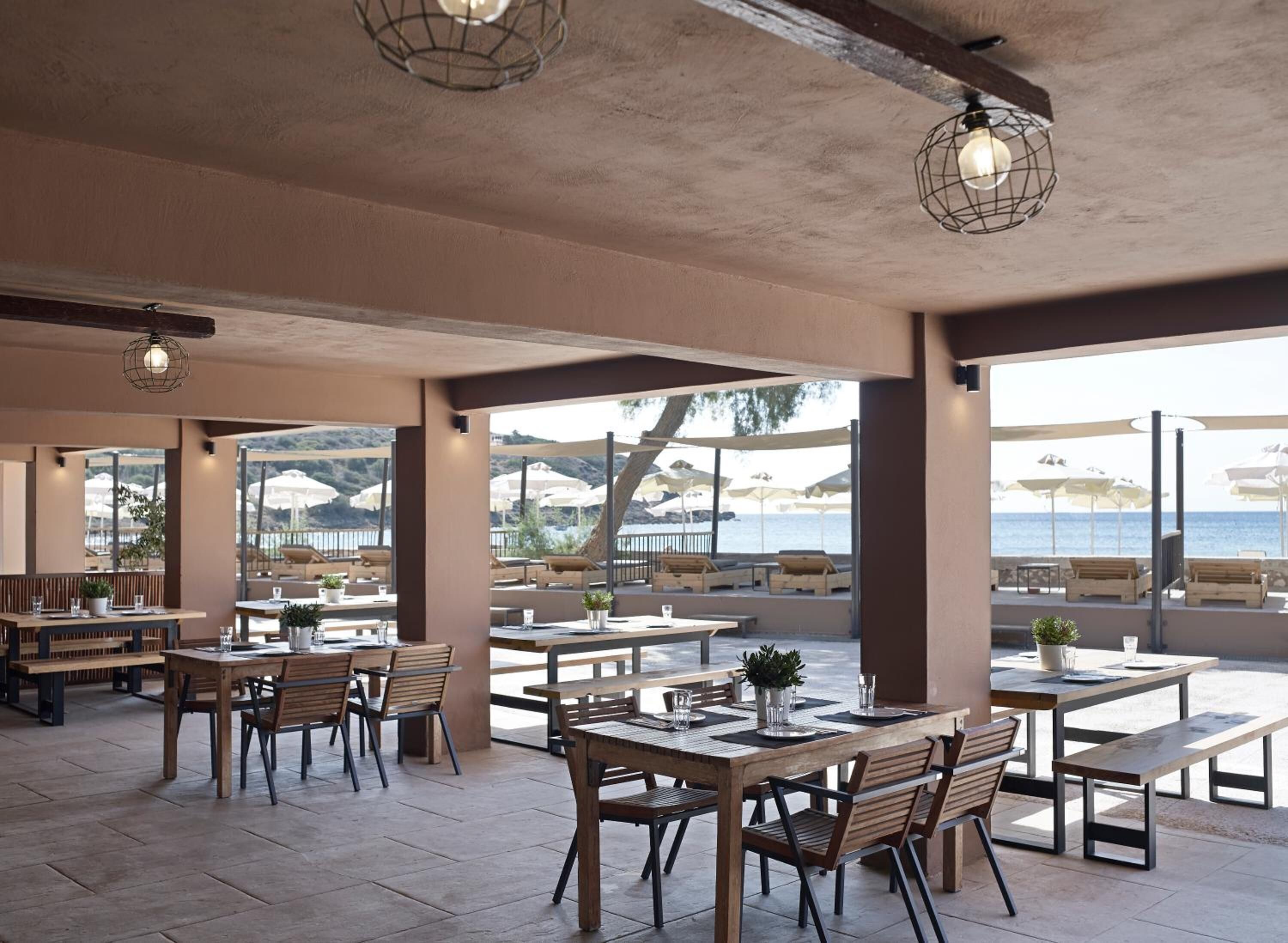 An outdoor dining area with wooden tables and chairs, large windows offering a view of the sea, and hanging light fixtures.