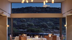 A dining area with tables set for four, large windows offering a view of a mountainous landscape, and pendant lighting. - Aegeon Beach Hotel (Lavreotiki)