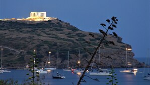 A coastal scene with a prominent white building on a hill, boats in the water, and a tree in the foreground. - Aegeon Beach Hotel (Lavreotiki)