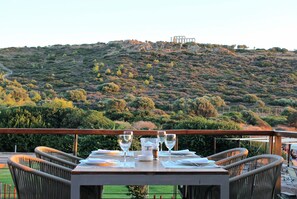 A dining table set outdoors with chairs, overlooking a hilly landscape with ruins. - Aegeon Beach Hotel (Lavreotiki)