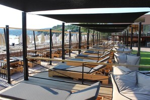 A beachside lounge area with sun loungers and umbrellas, wooden deck flooring, and a view of the sea. - Aegeon Beach Hotel (Lavreotiki)