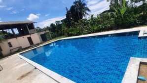 A swimming pool with blue tiles, surrounded by greenery and a building with a tiled roof.