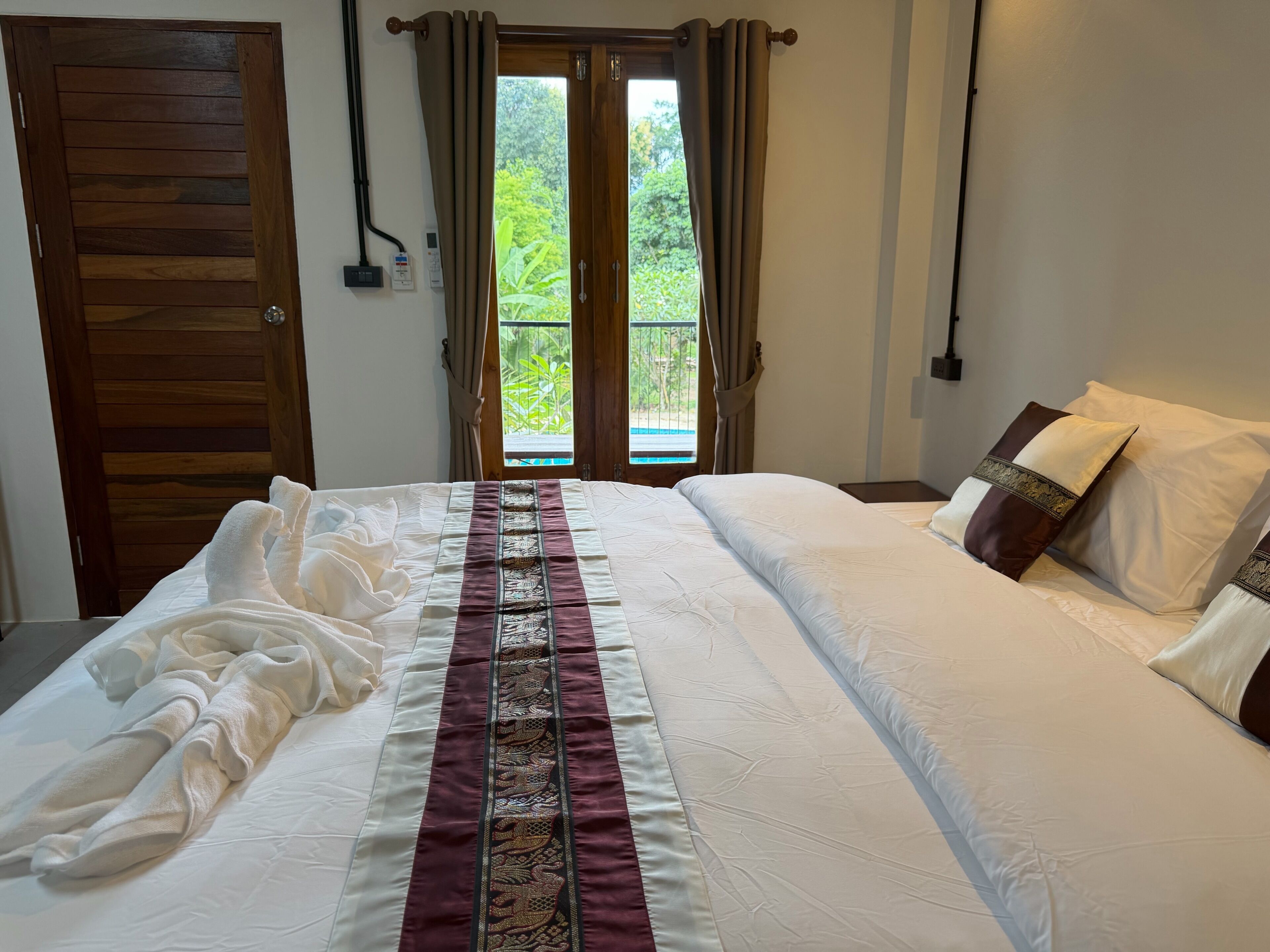 A hotel room with a bed, a window with curtains, and a view of greenery outside.