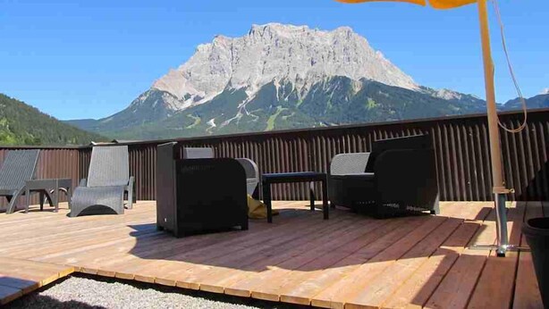 Terraza exterior con piso de madera, un paraguas amarillo y mobiliario para exteriores. Se ve una montaña al fondo.