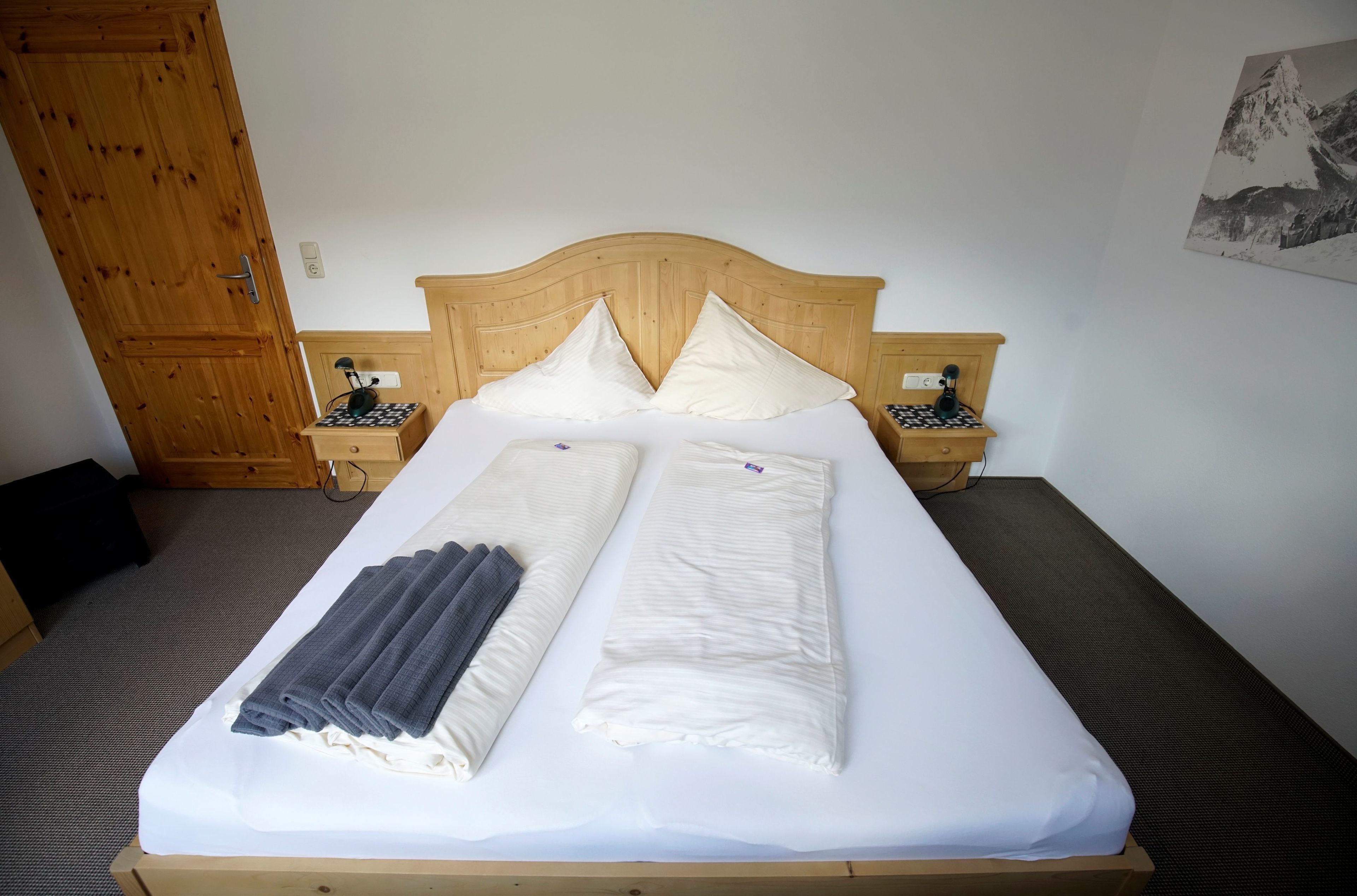 A neatly made bed with pillows and folded towels, wooden headboard, bedside tables with lamps, and a door in the background.