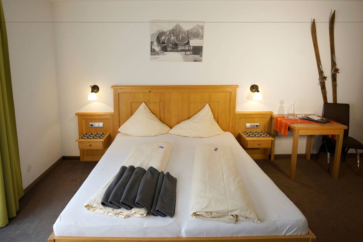 A neatly made bed with pillows and folded blankets, wooden headboard, bedside tables with lamps, a small table with a vase, and a framed picture on the wall.