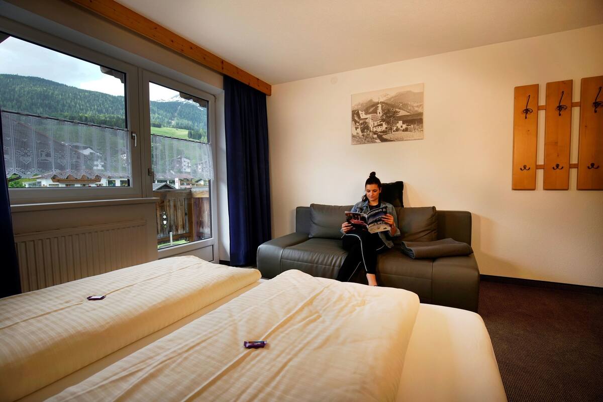 A hotel room with two beds, a sofa, a window with a view of mountains, and a wall-mounted coat rack.