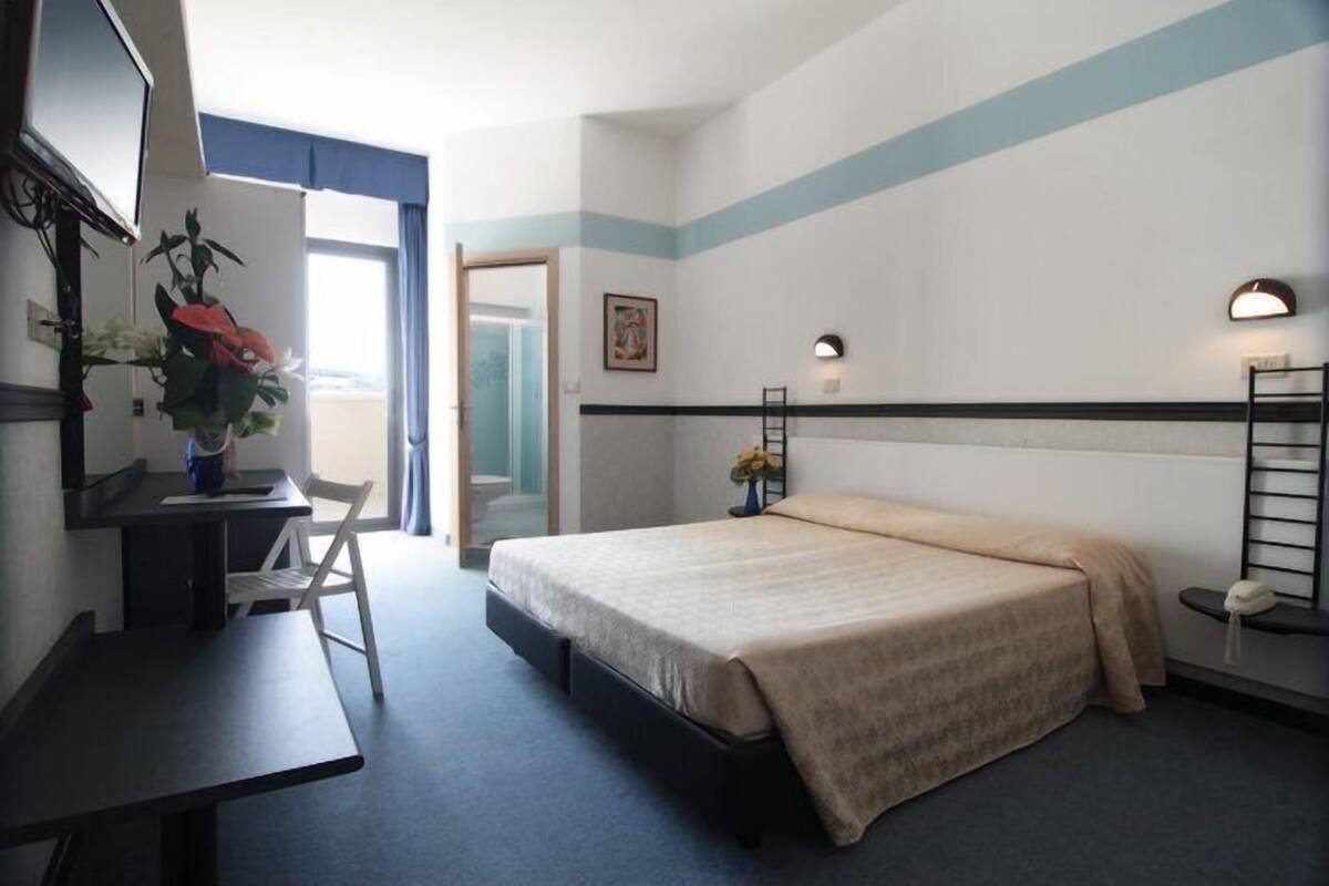Um quarto com cama, escrivaninha, cadeira e televisão.
