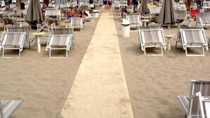 Zona de playa con filas de tumbonas y sombrillas, un paseo marítimo de arena y algunas personas a lo lejos.