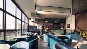 Un área de bar moderna con mesas redondas y sillas tapizadas en azul, ventanas amplias y una gran obra de arte en la pared.