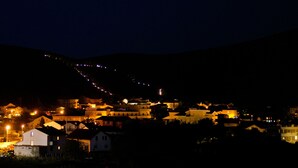 A nighttime view of a hillside village with illuminated buildings and a string of lights ascending the slope.
