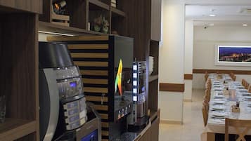 Un espace café moderne équipé de machines à expresso et d’une machine à café numérique, une longue table à manger dressée pour un repas, et un téléviseur fixé au mur.