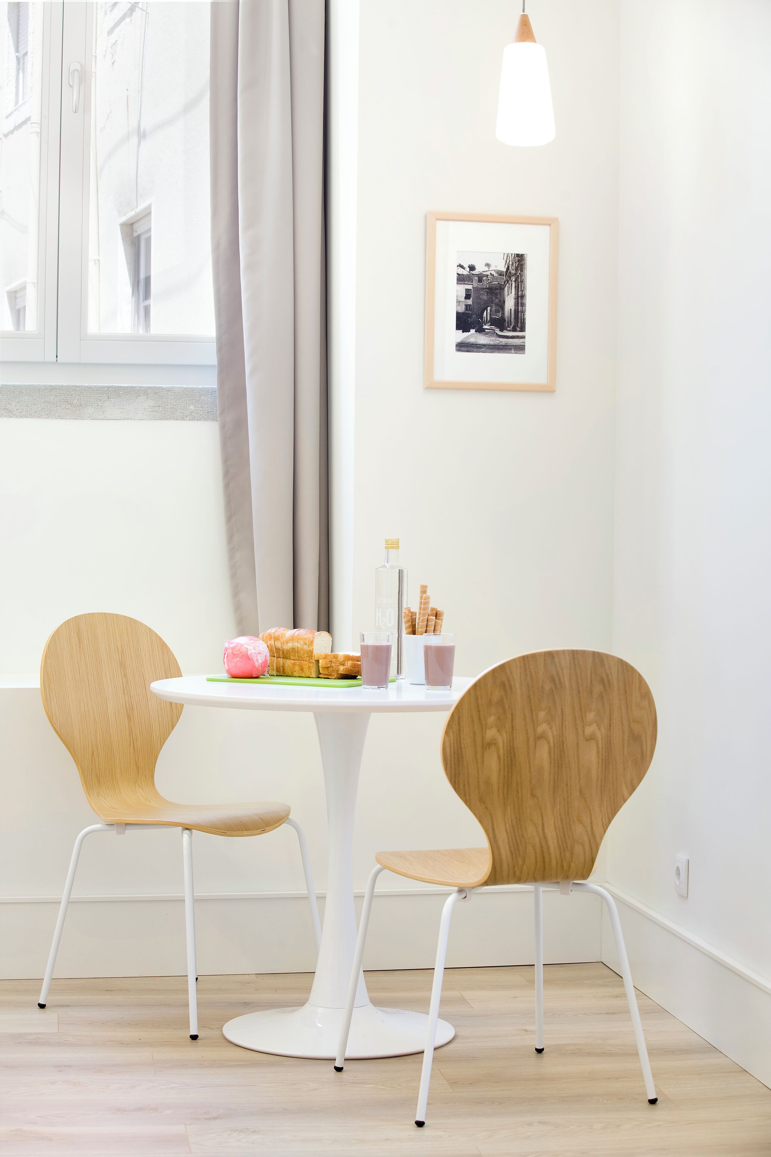 A room with a round table, two chairs, a framed picture on the wall, and a window with curtains.