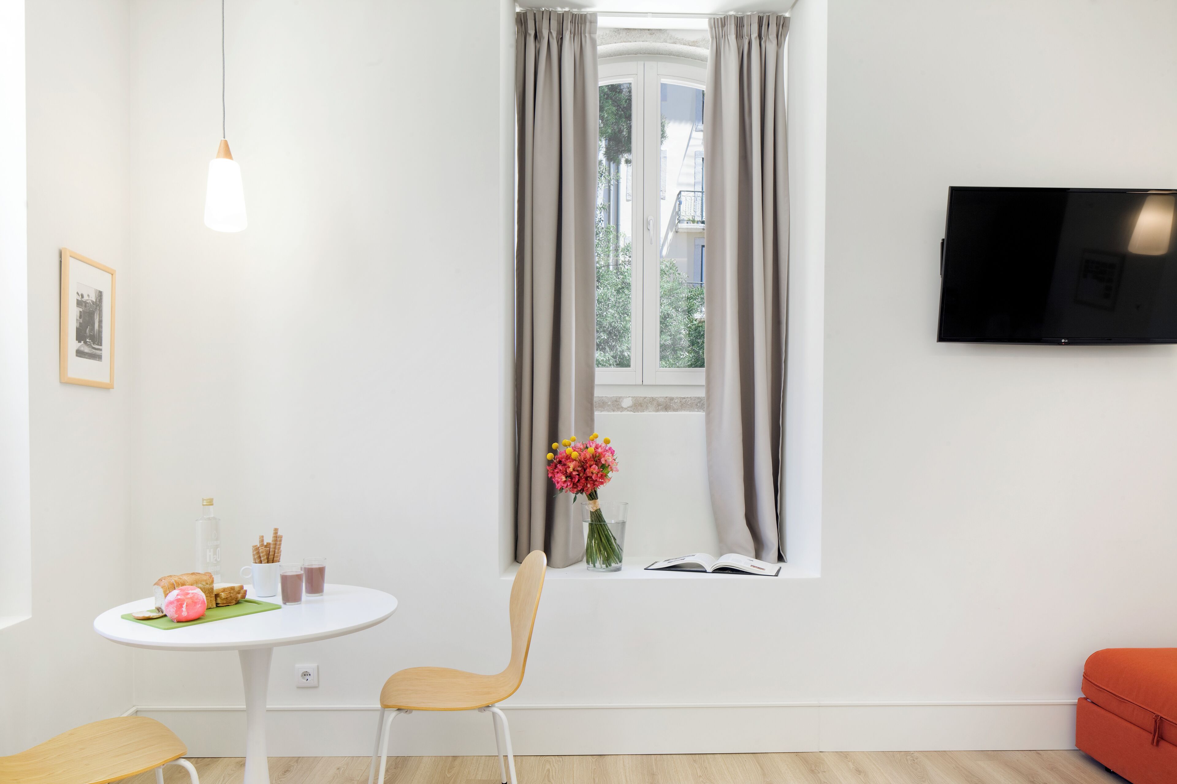 Ein modernes Zimmer mit einem weißen Tisch, einem Stuhl, einem an der Wand befestigten Fernseher und einem Fenster mit Vorhängen.