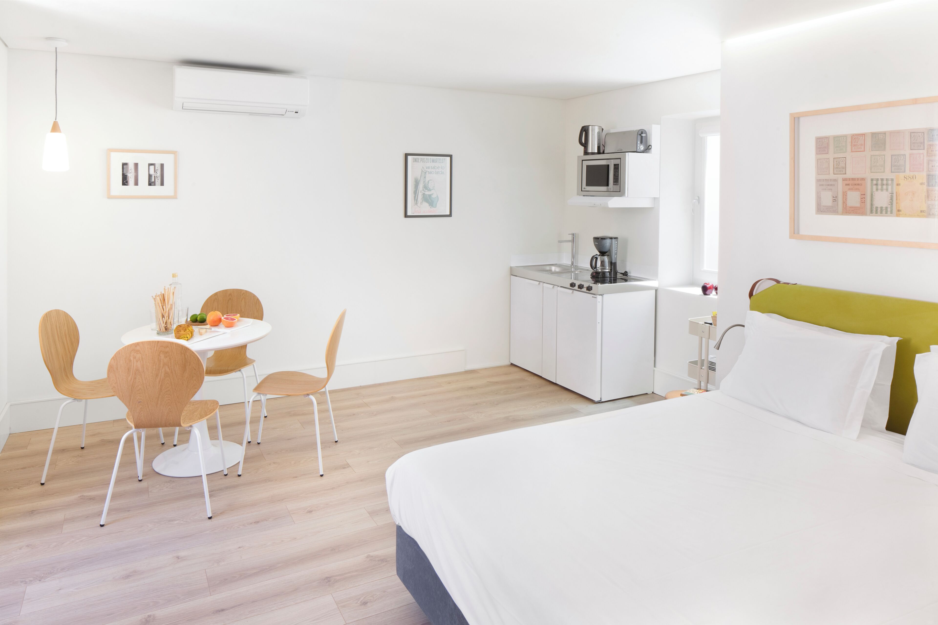 A modern hotel room with a bed, a dining table with chairs, a kitchenette, and a framed picture on the wall.