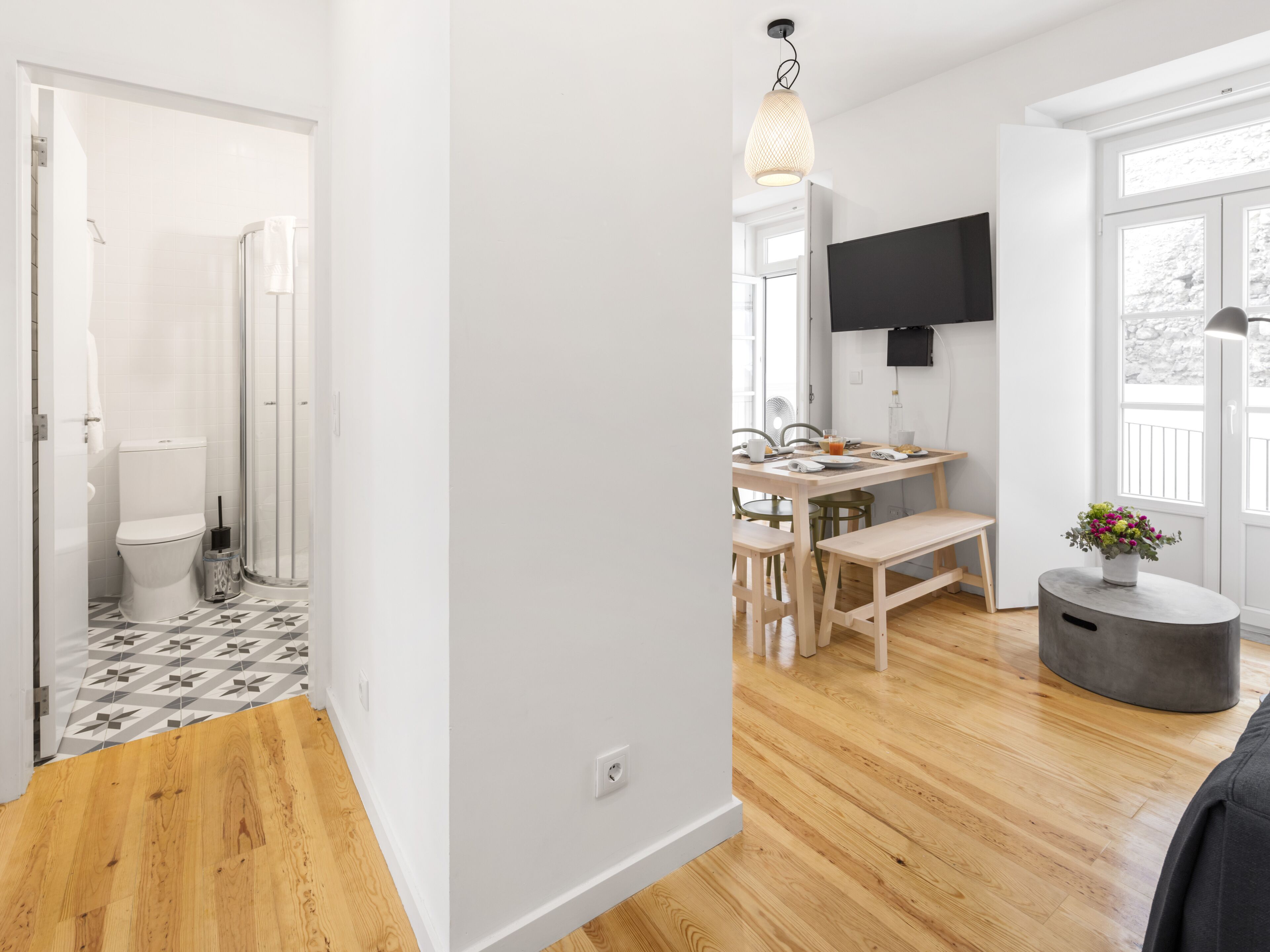 Un apartamento moderno con suelo de madera, una cocina con comedor, un televisor montado en la pared y un baño con ducha e inodoro.