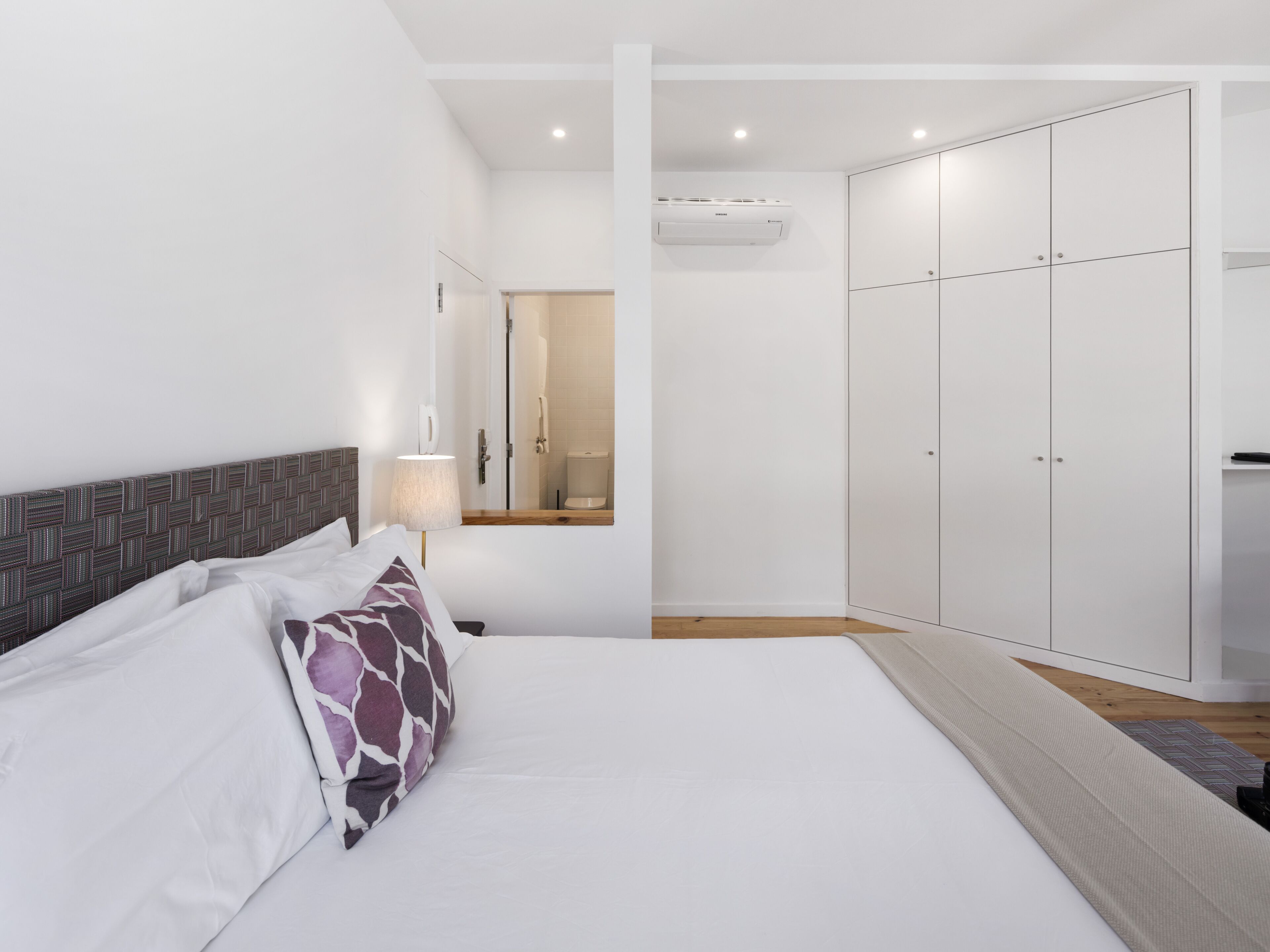 A bedroom with a bed, a nightstand, a lamp, a wardrobe, and a bathroom visible through an open door.