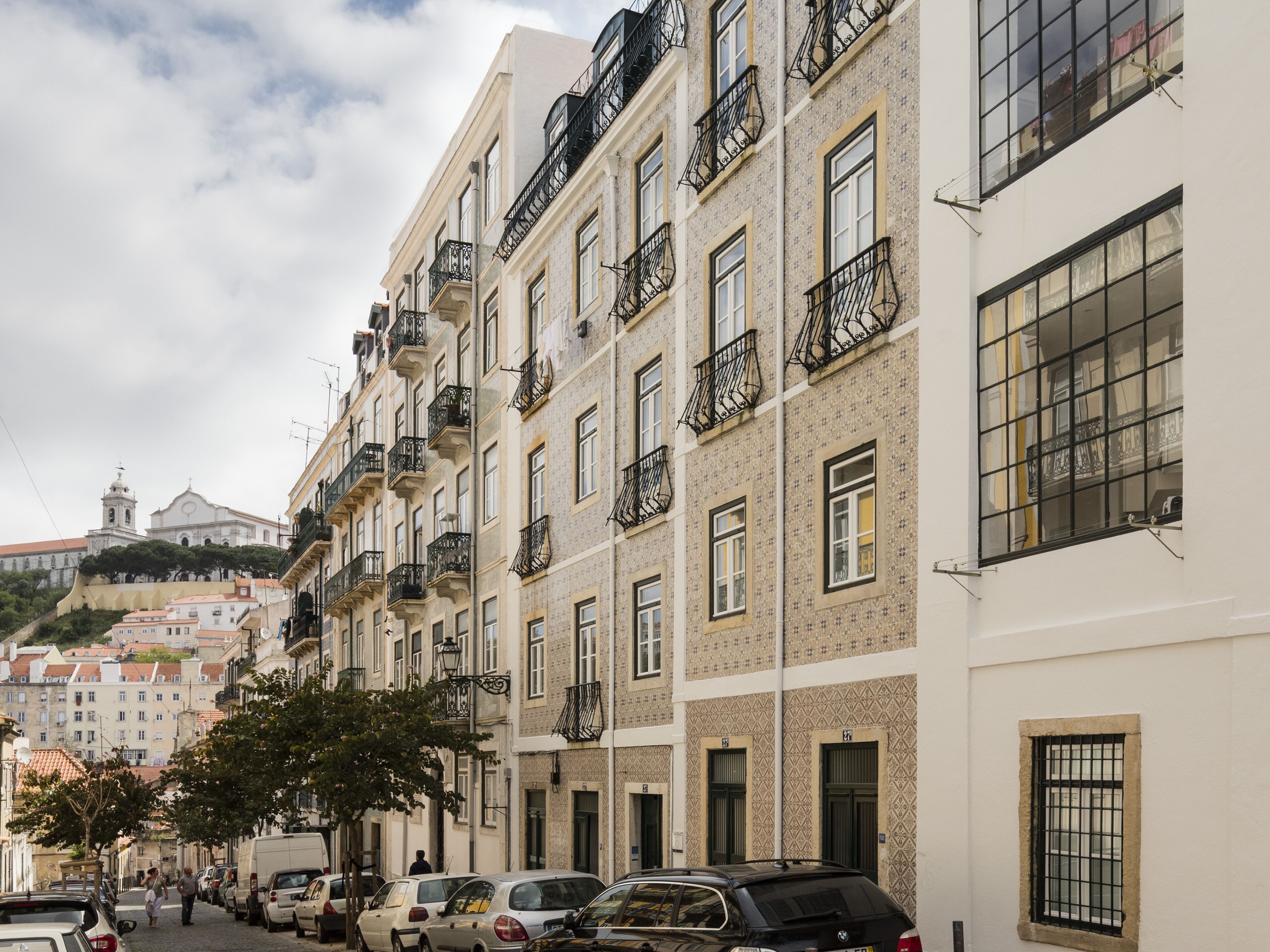 A row of multi-story buildings with balconies, parked cars, and a church in the background.