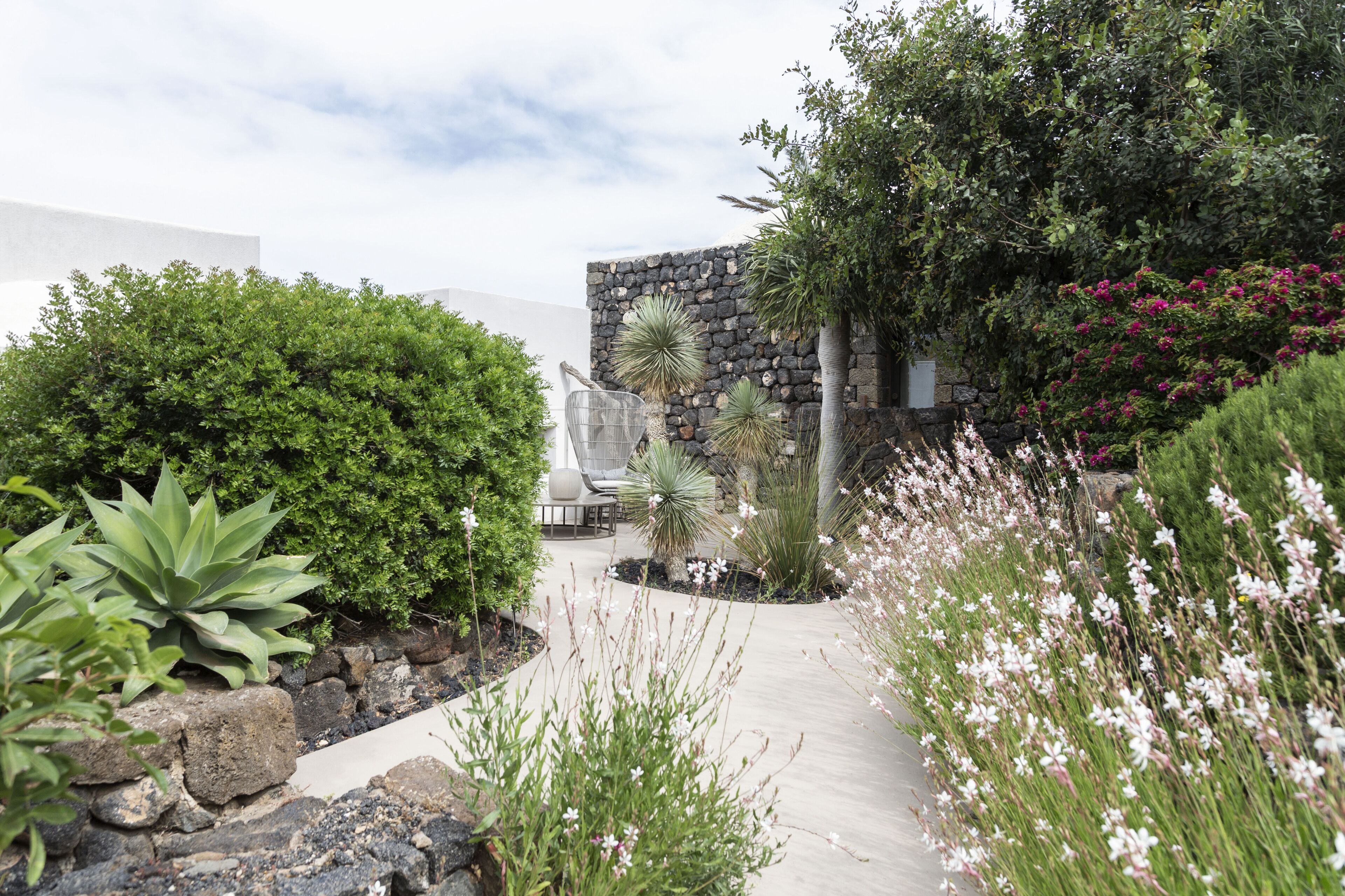 Un jardín ajardinado con diversas plantas, un muro de piedra y una zona de estar con una silla y una mesa.