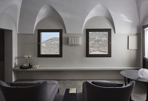 Une pièce moderne avec des plafonds en arc de cercle, deux fenêtres donnant sur un paysage vallonné, une petite table avec un livre et deux chaises de couleur sombre.