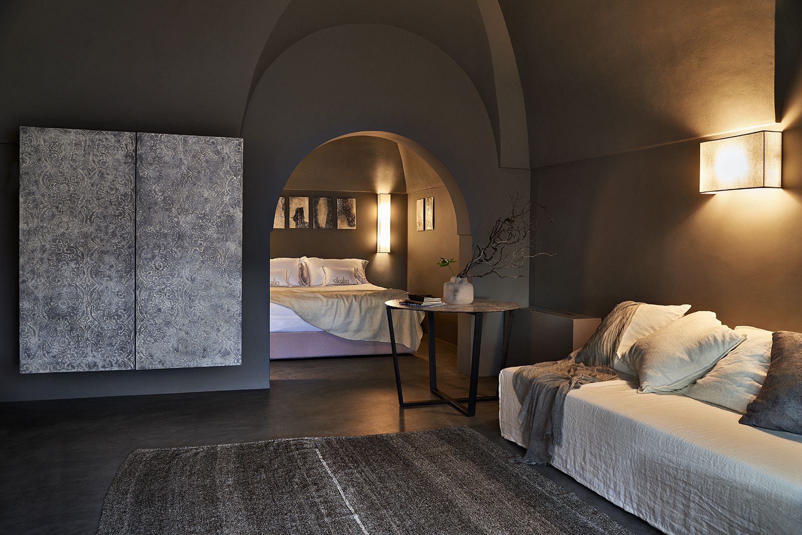 Een moderne slaapkamer met een groot bed, een zithoek met een bank, en een klein tafeltje. Aan de muur hangt een groot kunstwerk en op het tafeltje staat een decoratieve plant.