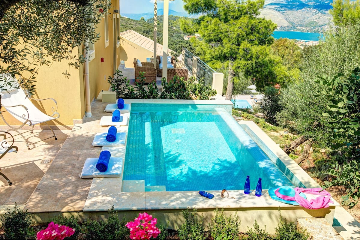 A pool area with a view of the sea, surrounded by greenery and a building with a yellow facade.