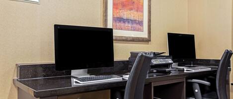 A business center with two computer stations, chairs, and a sign indicating it is open 24 hours.