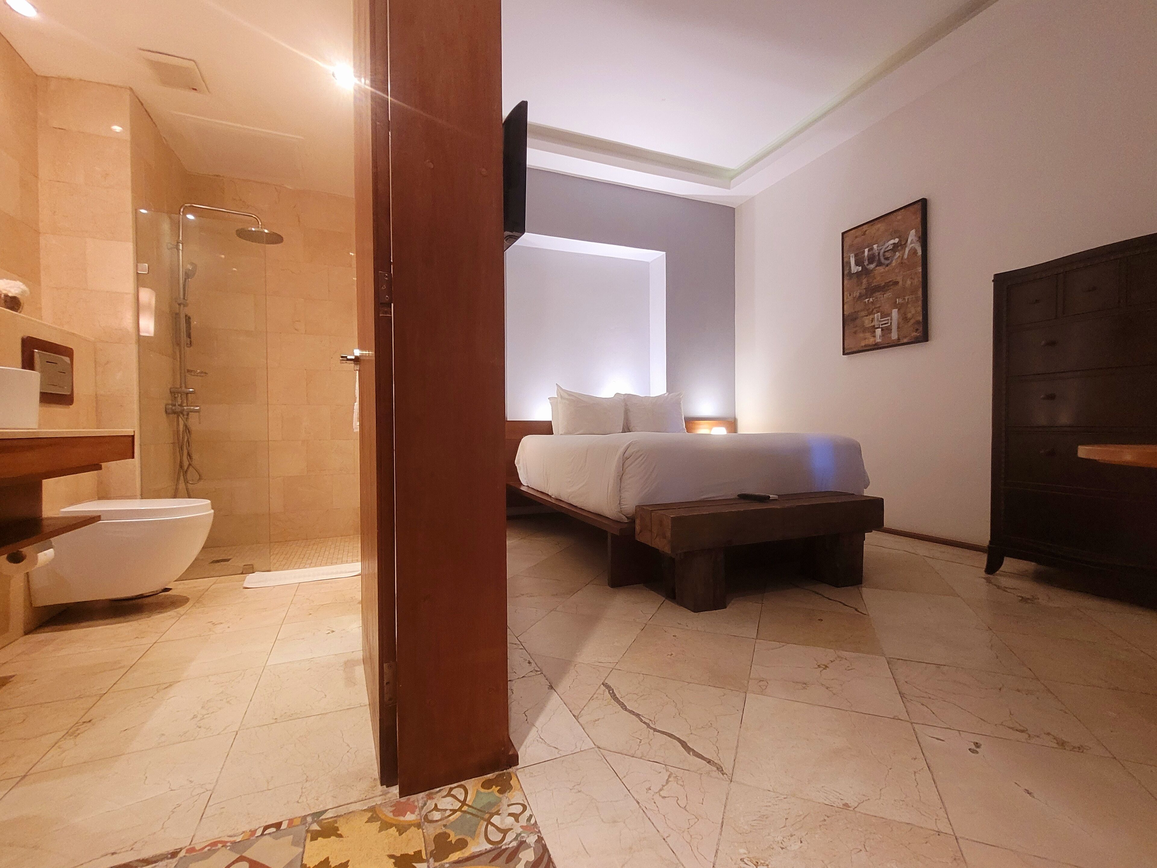 A modern bathroom with a shower, toilet, and sink. There is a bed with a nightstand and a wooden bench.