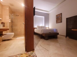 Uma casa de banho moderna com chuveiro, sanita e lavatório. Há uma cama com uma mesinha de cabeceira e um banco de madeira.