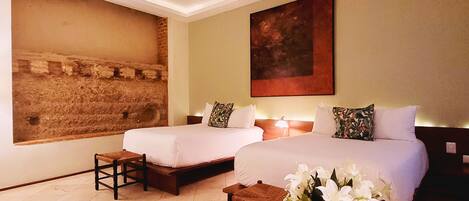 A hotel room with two beds, a wooden bench, a small table with a vase of flowers, and a large painting on the wall.