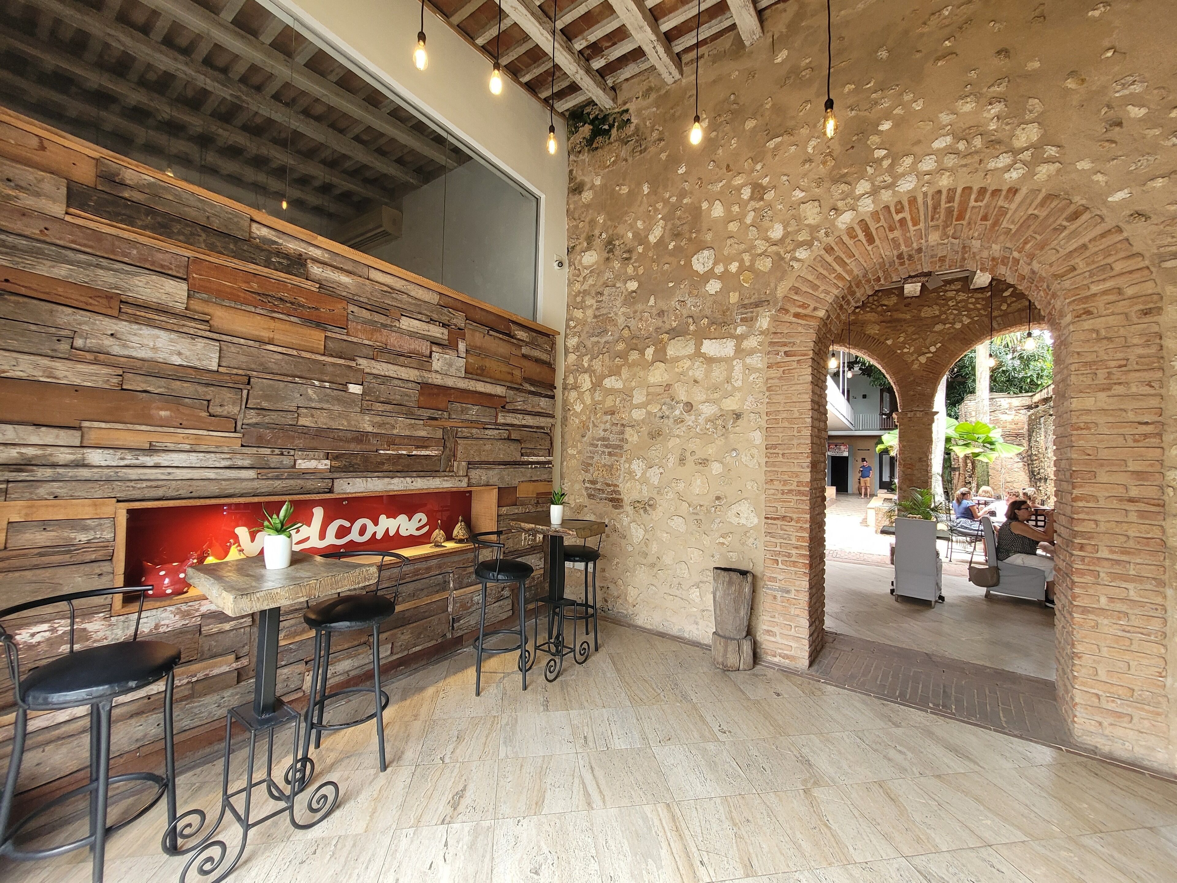 Una zona de bar con una pared de madera rústica, un cartel de 'Welcome' y un arco de piedra que da paso a un área de estar al aire libre.