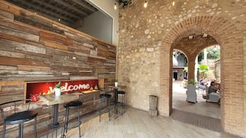 Una zona de bar con una pared de madera rústica, un cartel de 'Welcome' y un arco de piedra que da paso a un área de estar al aire libre.
