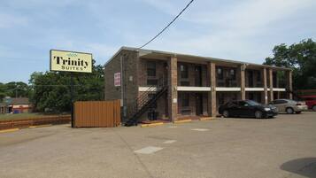 A brick building with a sign for Trinity Suites. It has a staircase leading to the entrance, a parking lot with cars, and a fenced area.