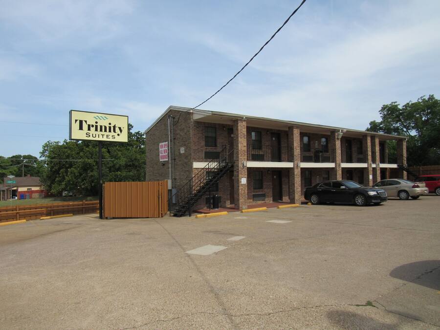 A brick building with a sign for Trinity Suites. It has a staircase leading to the entrance, a parking lot with cars, and a fenced area.