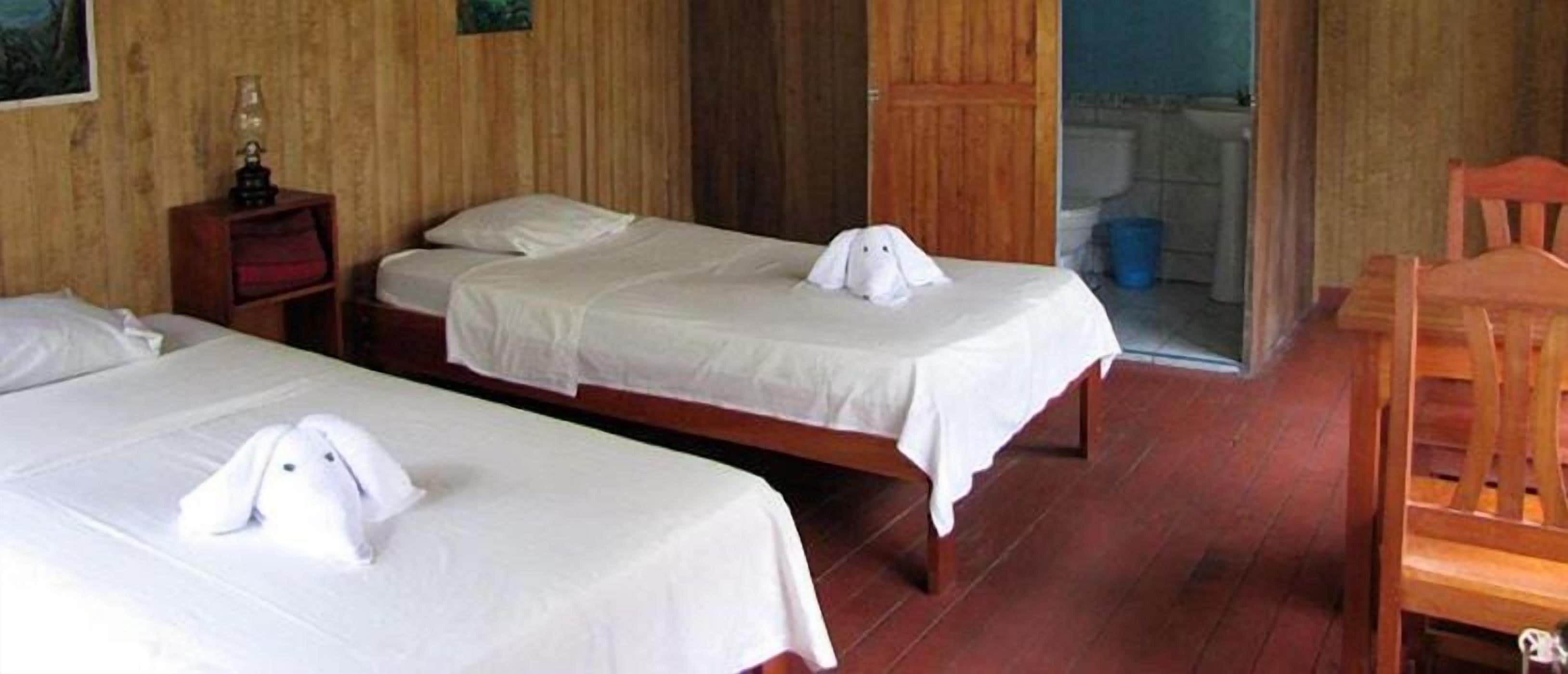A hotel room with two beds, a wooden floor, a wooden chair, and a small table with a lamp.