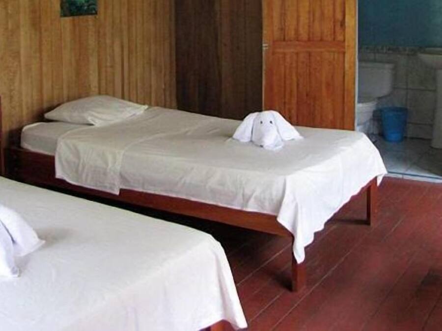 A hotel room with two beds, a wooden floor, a wooden chair, and a small table with a lamp.