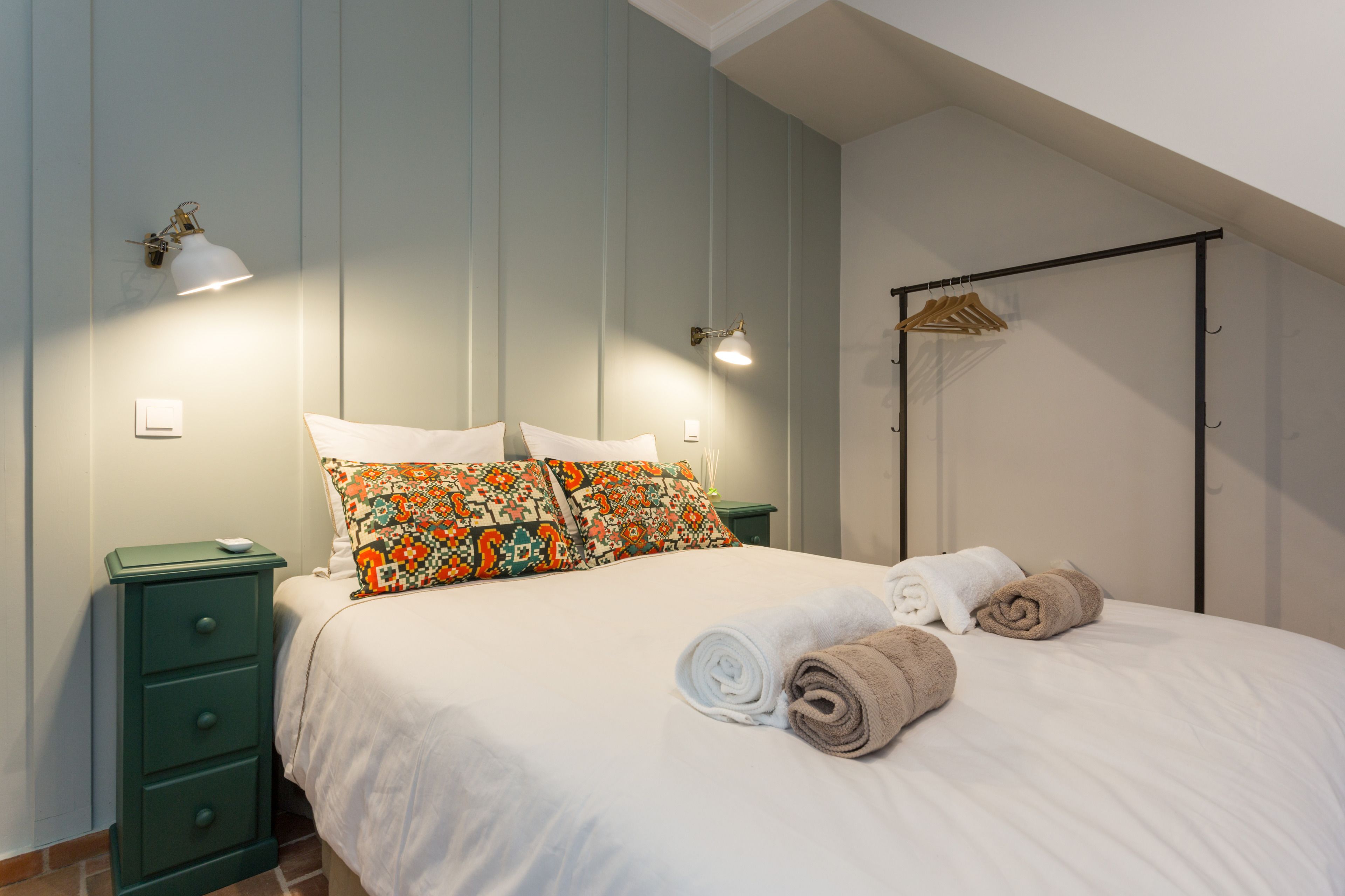 A neatly made bed with white and brown towels, a green nightstand with a lamp, and a wooden hanger rack against the wall.