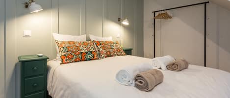 A neatly made bed with white and brown towels, a green nightstand with a lamp, and a wooden hanger rack against the wall.