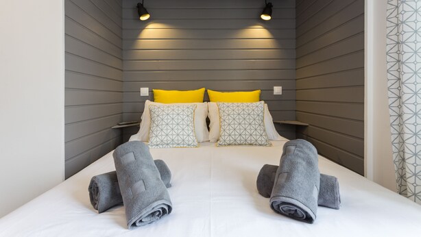 Un dormitorio moderno con pared gris, dos mesitas de noche, una cama con ropa blanca y dos almohadas amarillas, además de dos toallas grises.