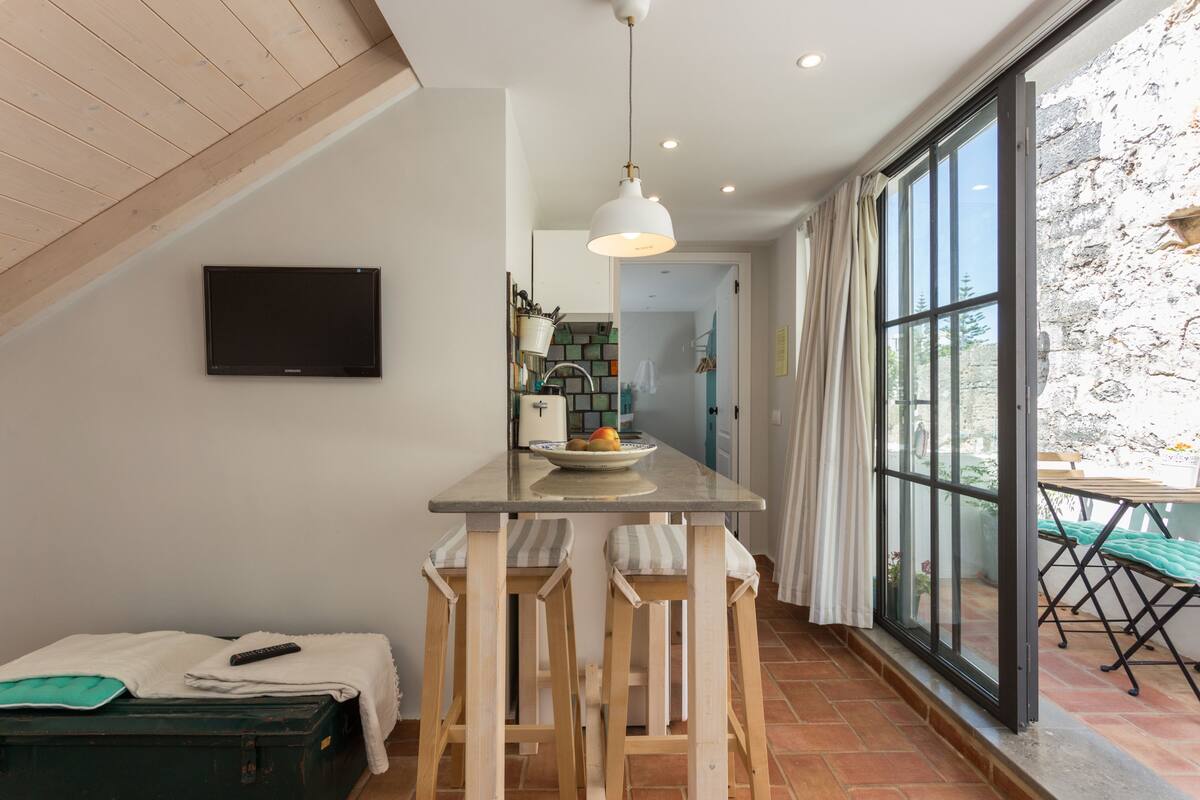 A modern kitchen with a large window, wooden stools, and a TV mounted on the wall.