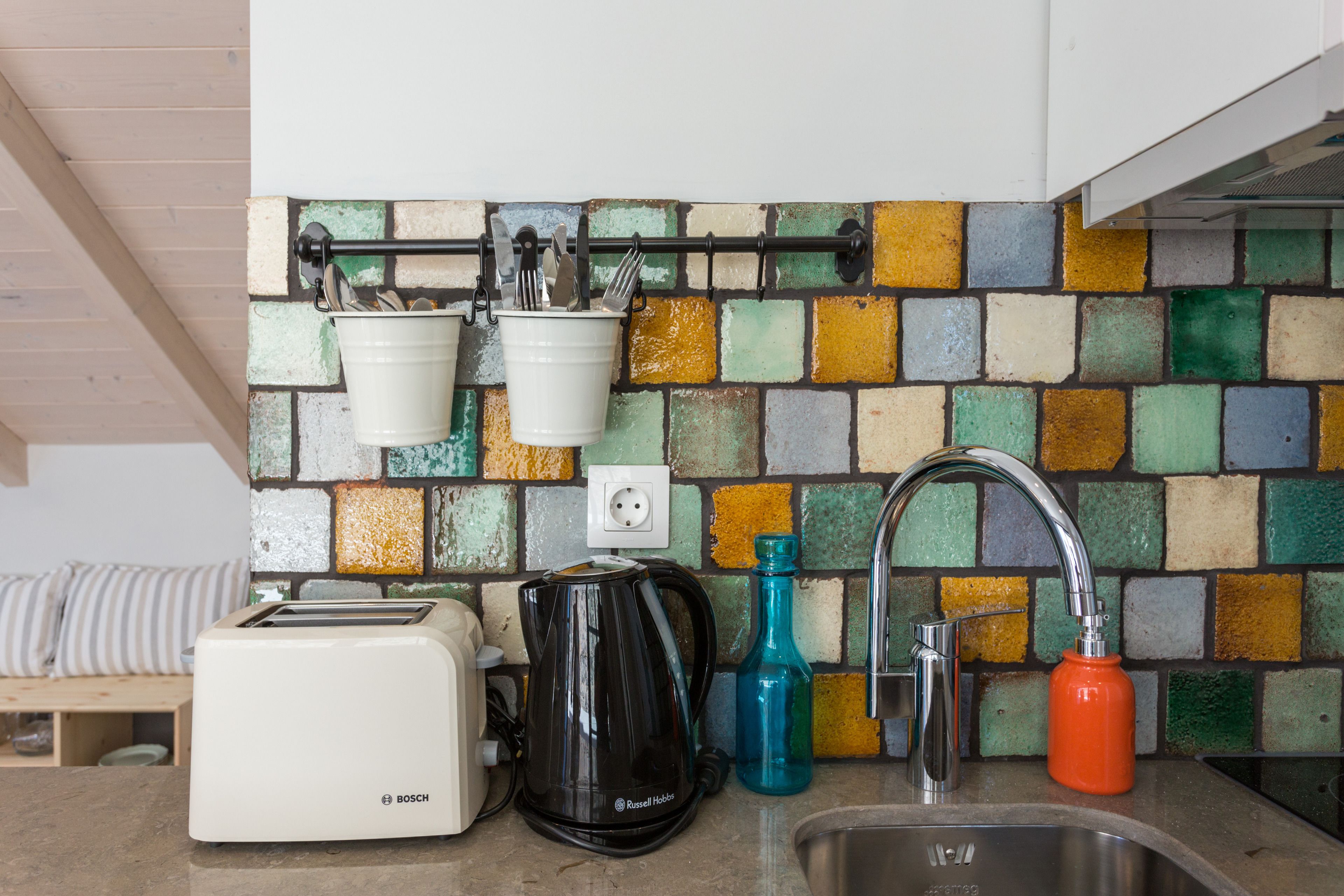 A kitchen with a colorful mosaic backsplash, a Bosch toaster, a Russell Hobbs kettle, a sink, and a faucet.