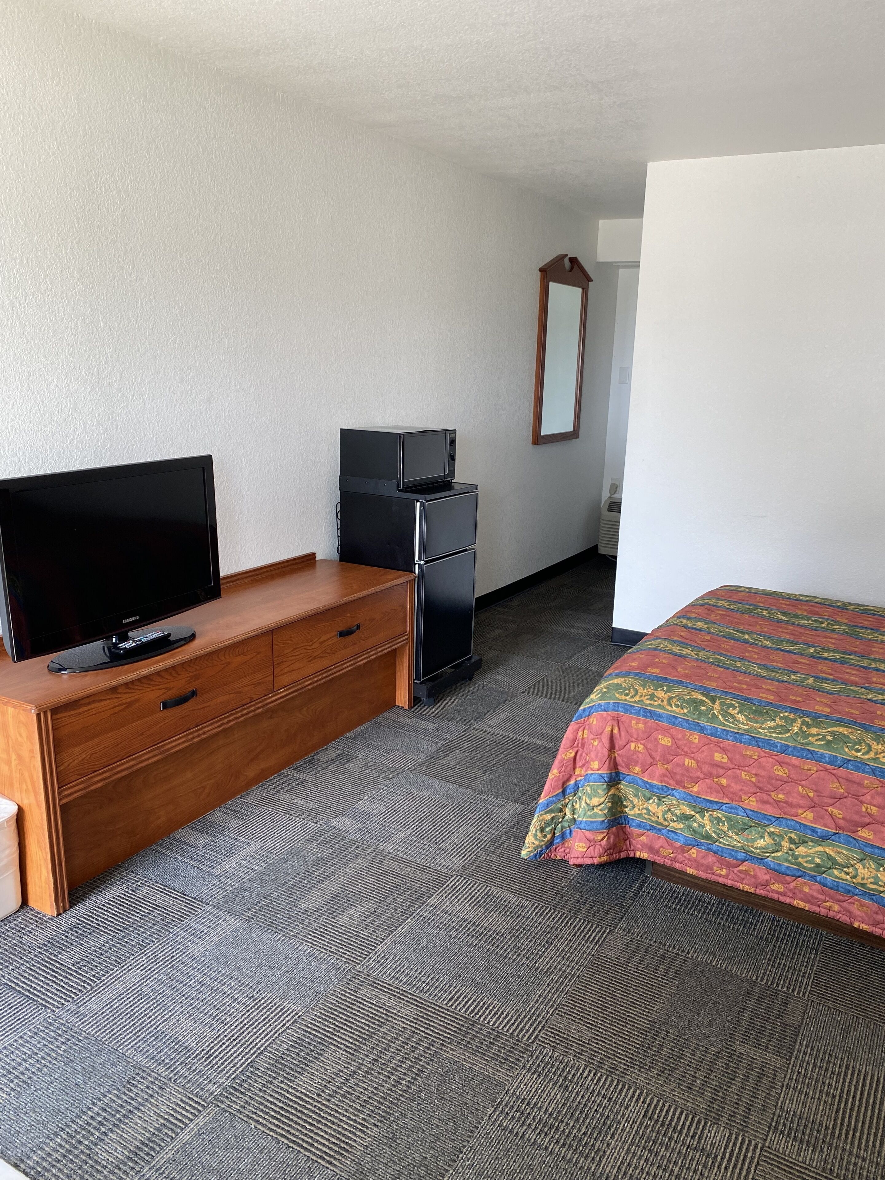 Ein Hotelzimmer mit einem hölzernen Schminktisch, einem Flachbildfernseher, einem kleinen schwarzen Kühlschrank, einem Bett mit einer farbenfrohen Steudecke und einem Spiegel an der Wand.