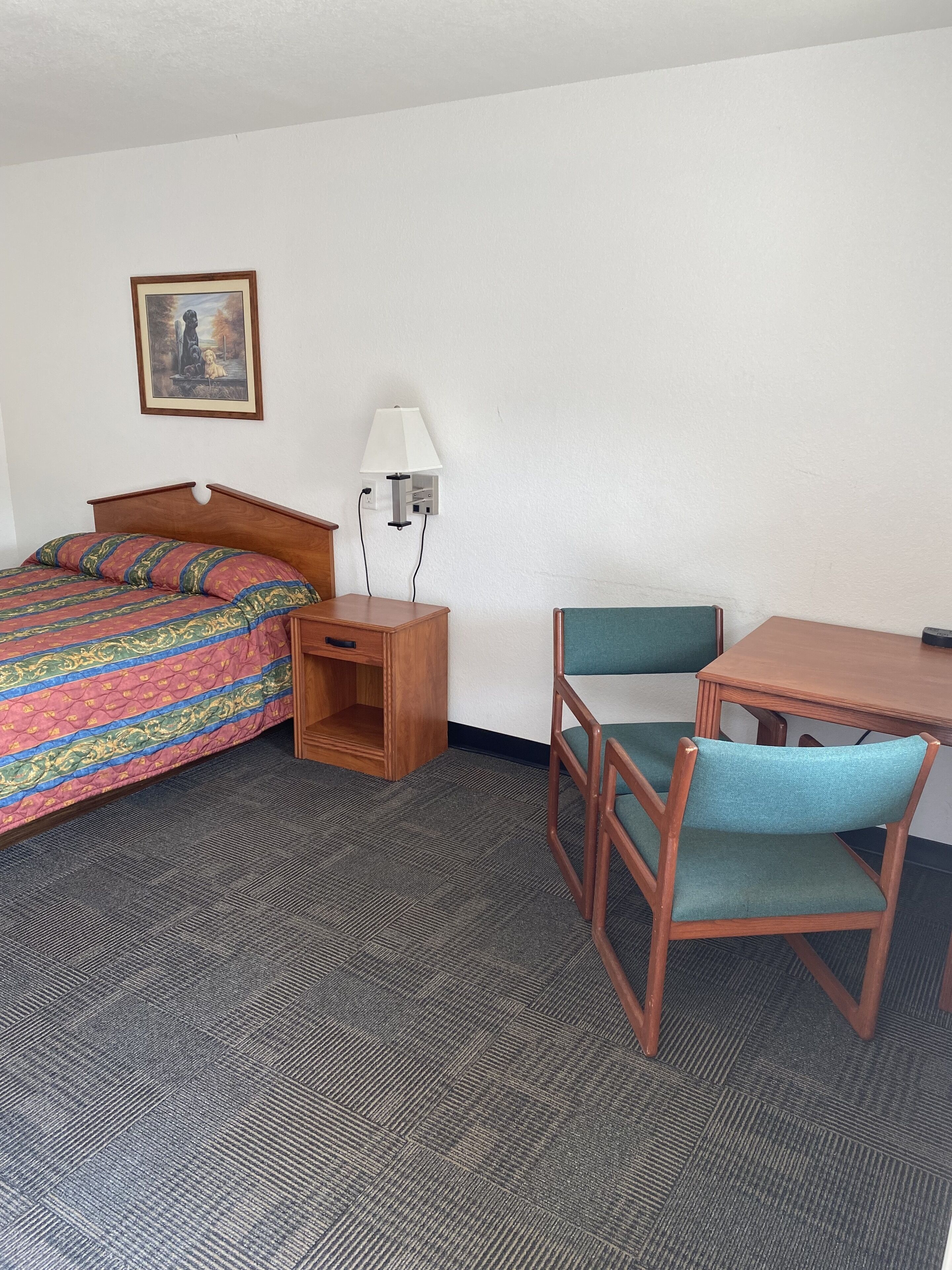 A hotel room with a bed, a desk, a chair, a lamp, and a painting on the wall.