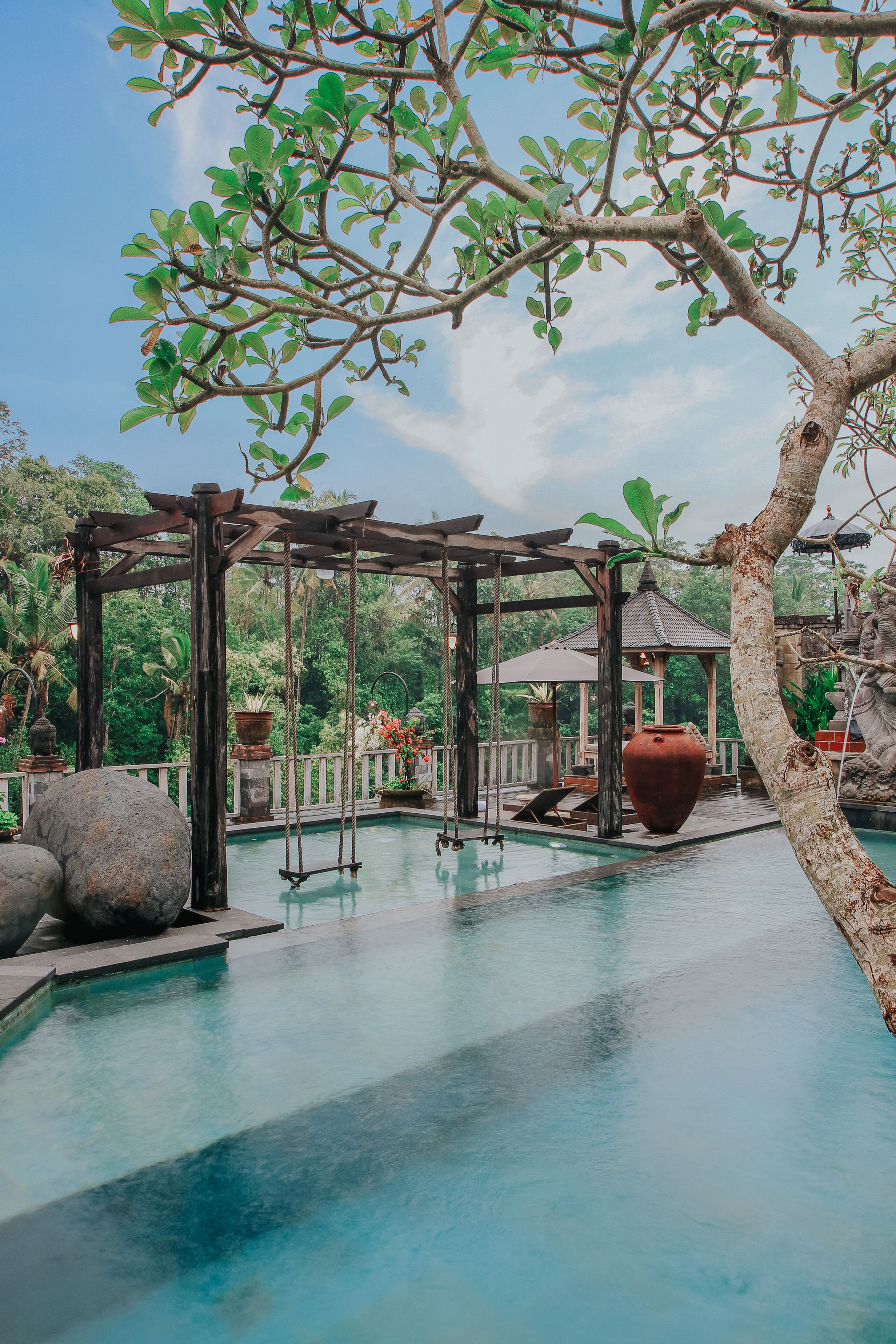 A serene outdoor pool area with a wooden swing, a gazebo, and lush greenery.
