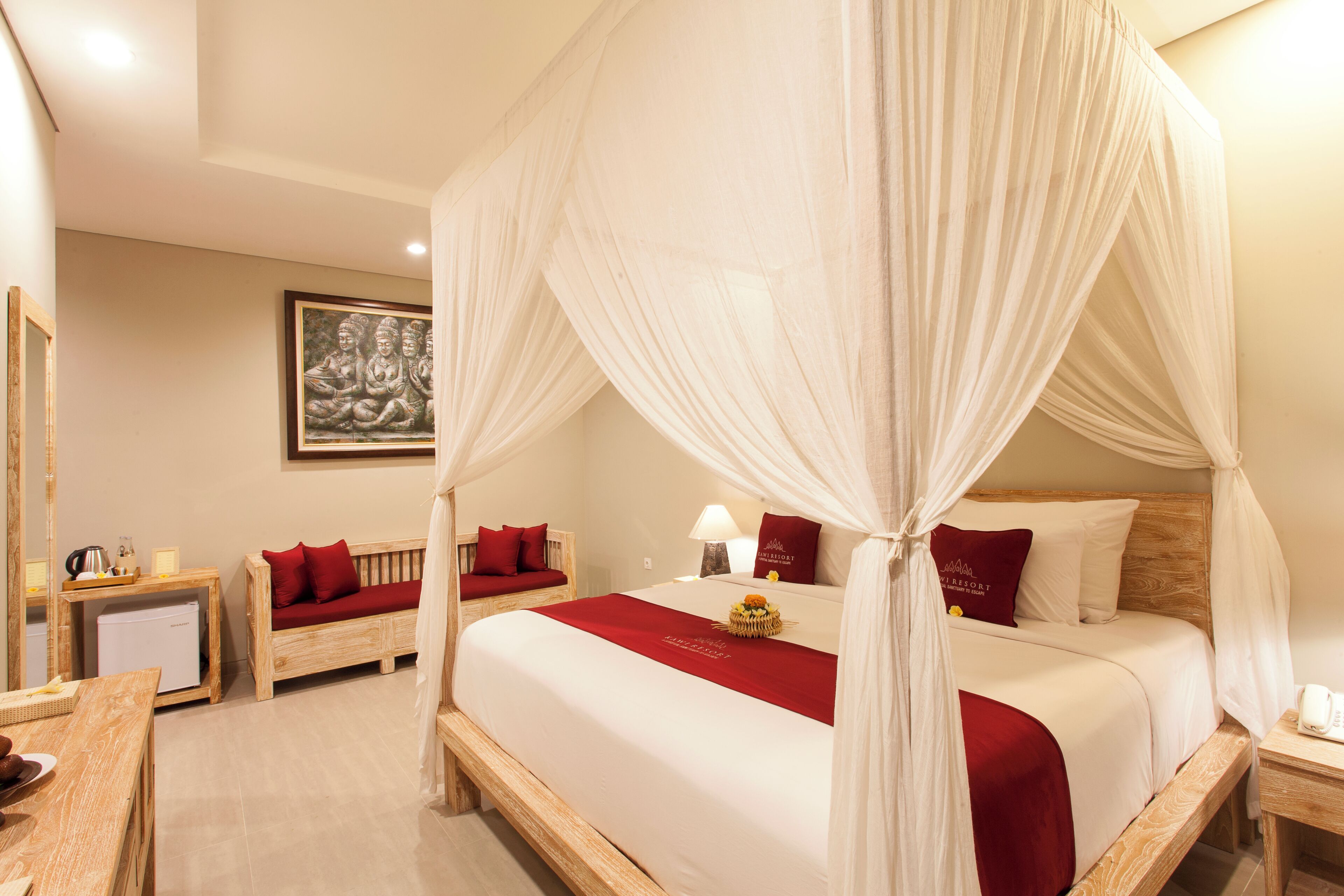 A hotel room with a four-poster bed, a wooden desk, a small refrigerator, and a painting on the wall.