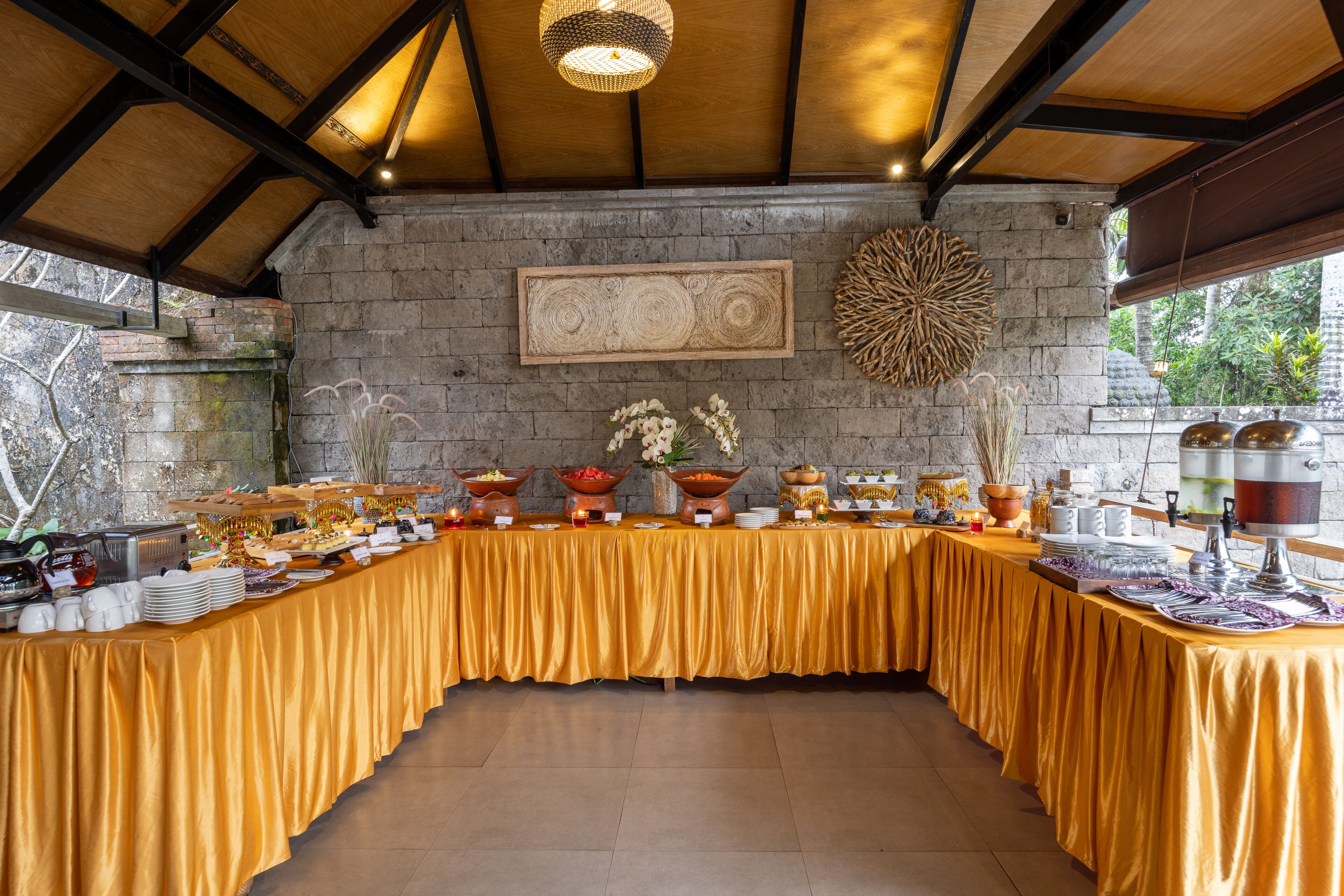 A buffet setup with a variety of food items and decorative elements on a long table, set against a stone wall with a wooden ceiling.