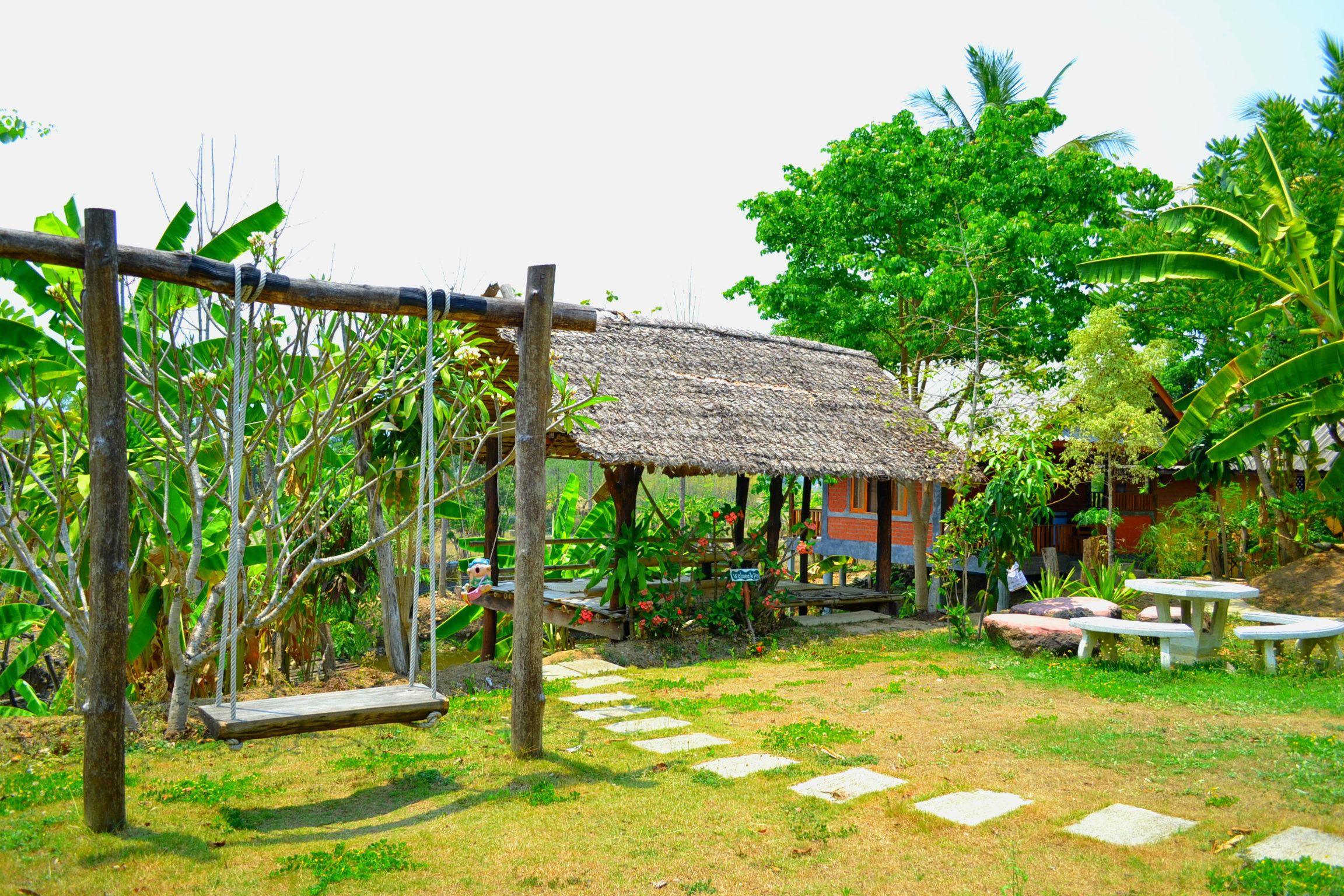 A rustic outdoor area with a swing set, a thatched-roof structure, and a stone pathway.